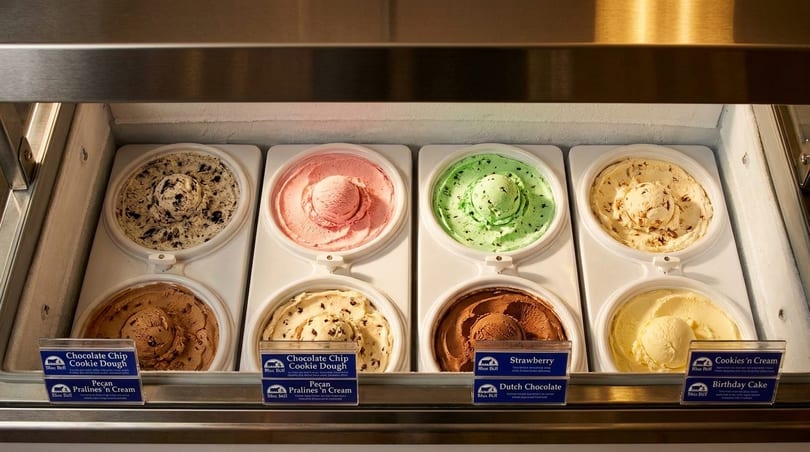 Colorful ice cream flavors with scoops in a refrigerated display case, with flavor labels.