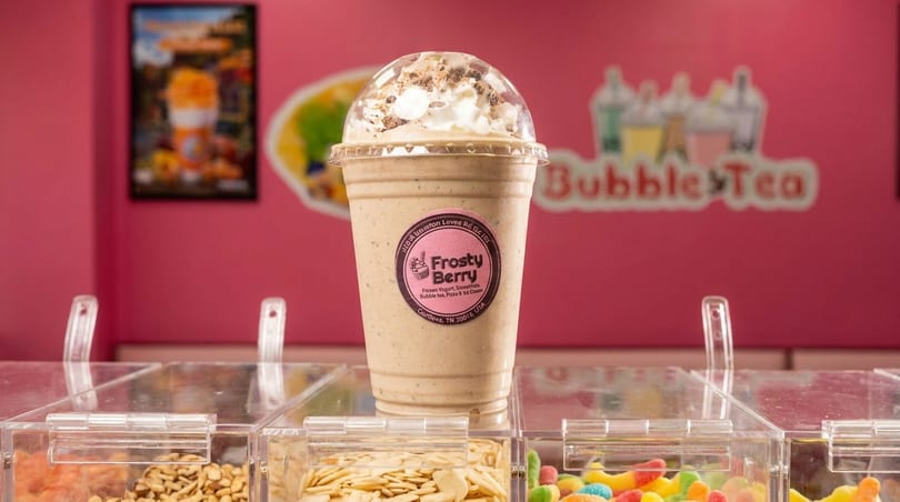 Frosty Berry drink with whipped cream and toppings on a counter with candy, against a pink "Bubble Tea" wall.